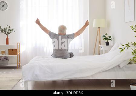 Uomo anziano che indossa indumenti da sonno che si sveglia a letto e si allunga le braccia seduto nella camera da letto di casa Foto Stock
