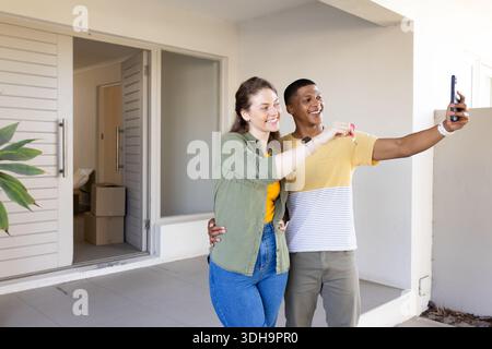 Coppia diversificata che scatta selfie con lo smartphone tenendo le chiavi della casa vicino alle scatole in movimento all'ingresso Foto Stock
