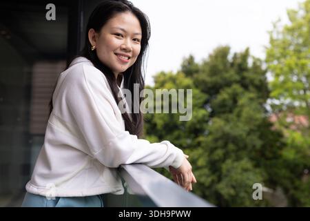 Donna asiatica in piedi sul balcone appoggiata su una guida di vetro sorridente indossando canottiere, spazio copia Foto Stock
