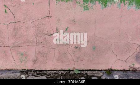 Primo piano dettagliato di una parete rosa intemprata caratterizzata da numerose crepe, vernice che sbucciava gli strati verdi e una texture ruvida e invecchiata sopra il gro scuro Foto Stock