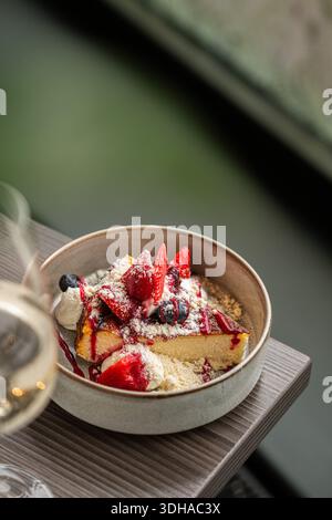 Una deliziosa fetta di cremosa cheesecake basca bruciata servita in un recipiente di ceramica, condita con fragole fresche, mirtilli e zucchero a velo. Foto Stock