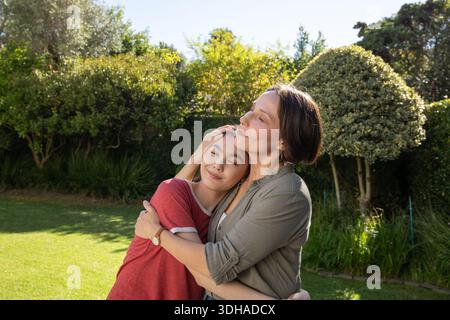 Madre e figlia asiatica si abbracciano e si appoggiano nel cortile, mostrando orologio da polso e orecchini a cerchio. Abbracciare, stile di vita, candidi, stock, giardino, serenità, bon Foto Stock