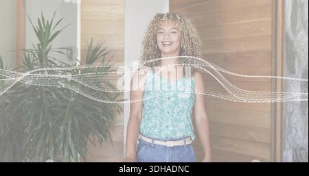 Donna sorridente che indossa un top floreale color ottanio e jeans in posa con una pianta e una grafica a onde punteggiate Foto Stock