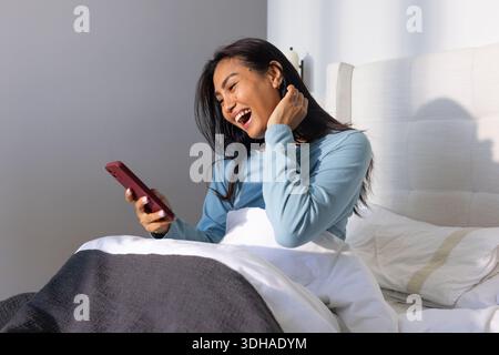 Donna asiatica seduta sul letto con la parte superiore blu, con uno smartphone con involucro rosso e ridendo Foto Stock