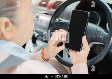 Vista posteriore di una donna matura con apparecchio acustico che utilizza il telefono cellulare per guardare la mappa di navigazione durante il viaggio in auto. Focalizzazione selettiva sul telefono. Foto Stock