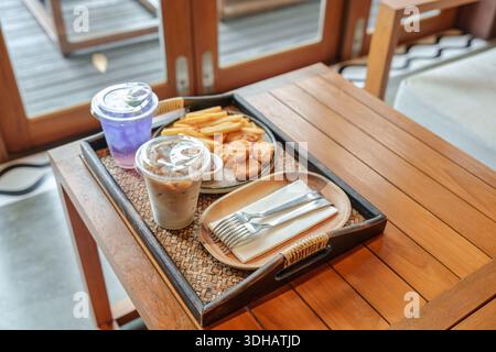Il vassoio contiene patatine fritte croccanti, pepite di pollo, caffè a latte freddo, rinfrescante bevanda di piselli farfalla servita su un tavolo di legno nell'accogliente caffè Foto Stock