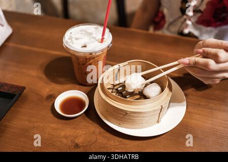 A mano con bacchette che raccolgono dim sum nel piroscafo di bambù e tè tailandese freddo su un tavolo di legno Foto Stock