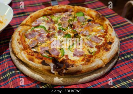 Pizza cotta al forno condita con prosciutto, peperone verde e formaggio con mealt servita in piatto su tovaglia di tartan Foto Stock