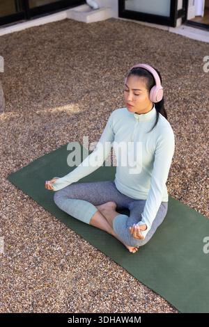 Donna asiatica che meditava sul tappeto verde sul cortile di ghiaia, con pullover alla menta e cuffie rosa Foto Stock