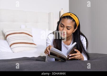 Donna asiatica sdraiata in camera da letto sul letto leggendo un libro in brossura aperto con fascia gialla, spazio per le copie Foto Stock