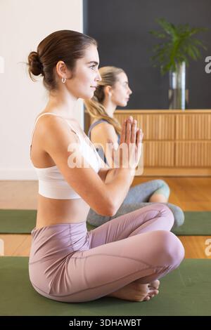 Due donne che meditano, sedute su tappeti verdi in studio con armadietto e pianta, indossando abbigliamento sportivo Foto Stock