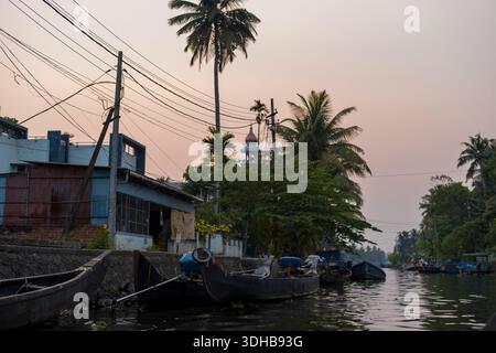 Alappuzha, India - 7 marzo 2023: Diverse barche di legno sono ormeggiate lungo un canale delimitato da mura di pietra, con case residenziali, palme, pali elettrici e cavi sospesi visibili al crepuscolo. Foto Stock