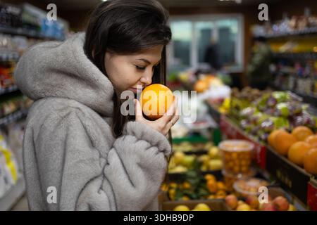 Una donna si trova in un mercato con un arancio vicino alla faccia. Lei odora la frutta e sembra premurosa. Lo sfondo ha molti tipi di frutta e verdura. Foto Stock