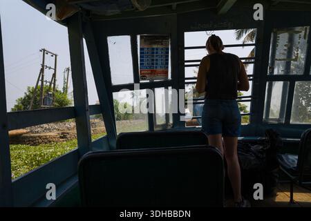 Alappuzha, India - 9 marzo 2023: Una persona si trova vicino alla finestra all'interno di un traghetto locale sulle acque secondarie, con un cartello di programma visibile sul muro e sullo scenario all'aperto, tra cui linee elettriche e vegetazione vista attraverso le finestre. Foto Stock