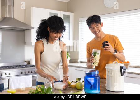 Diverse coppie che tritano cetrioli e preparano frullati all'isola della cucina, al bicchiere del frullatore e al telefono Foto Stock