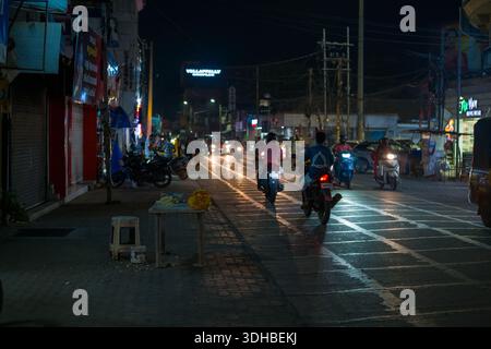 Alappuzha, India - 8 marzo 2023: Diverse motociclette e scooter viaggiano lungo una strada ben illuminata della città di notte, con vetrine illuminate, veicoli parcheggiati e un tavolino con ghirlande di fiori visibili sul marciapiede. Foto Stock