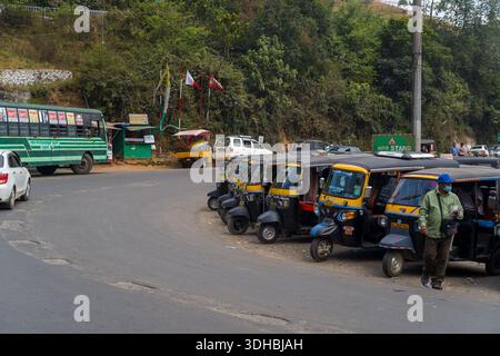 Munnar, India - 11 marzo 2023: Diversi risciò auto sono parcheggiati in un'auto accanto a una strada nel centro di Munnar, con un uomo che indossa una maschera davanti e un autobus pubblico verde visibile sullo sfondo. Foto Stock