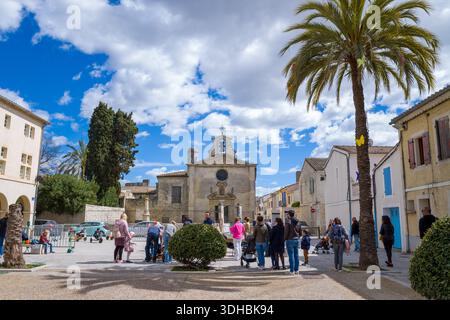 Luogo sconosciuto - 2 aprile 2023: Gruppi di persone si alzano e camminano in una piazza pubblica con palme, una moderna fontana asciutta e la storica Chapelle des Penitents Gris visibile sullo sfondo sotto un cielo parzialmente nuvoloso. Foto Stock