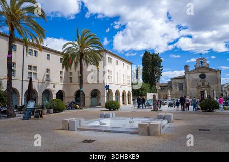 Posizione sconosciuta - 2 aprile 2023: Persone in piedi e camminare in una piazza pubblica con palme, una moderna fontana asciutta e la storica Chapelle des Penitents Gris visibile sullo sfondo sotto un cielo parzialmente nuvoloso. Foto Stock
