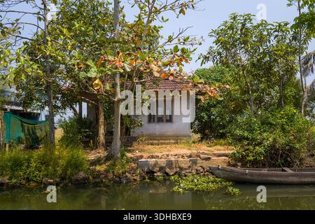Alappuzha, India - 9 marzo 2023: Una piccola casa con un tetto piastrellato è situata sulla riva delle backwaters, circondata da alberi e vegetazione, con una canoa in legno ormeggiata sul bordo dell'acqua. Foto Stock