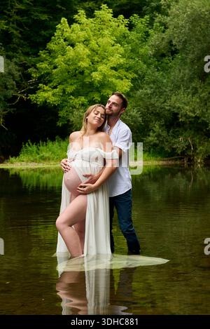 Donna incinta e il suo partner che si abbracciano teneramente nel fiume poco profondo nella foresta Foto Stock