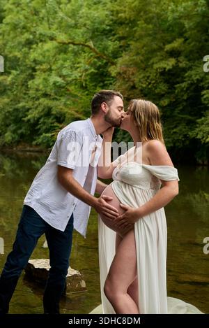 Aspettatevi una coppia che bacia e tocca la pancia incinta all'aperto in natura Foto Stock