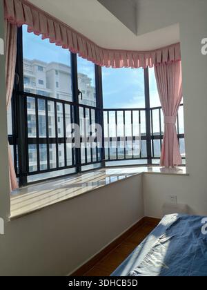 Accogliente camera da letto con finestra a bovindo e tende rosa che si affacciano sugli edifici urbani Foto Stock