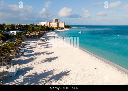 Le onde si infrangono dolcemente contro la riva di Noord, Aruba, dove una splendida spiaggia invita i visitatori a rilassarsi sotto le palme ondeggianti. Ideale per le vacanze, questo paradiso tropicale offre una fuga al sole e al mare. Foto Stock