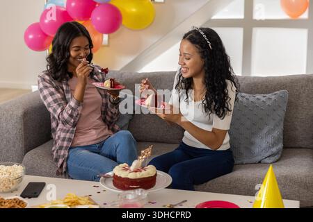 Diverse amiche sedevano sul divano grigio nel soggiorno mangiando una torta di velluto rosso con le candele. Celebrazione, amicizia, casalinga, accogliente, gioiosa, social, colorato || modello rilasciato Foto Stock