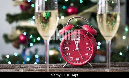 Due bicchieri pieni di bibita frizzante sedetevi vicino a un orologio rosso su un tavolo di legno. Un albero di Natale decorato con luci colorate e ornamenti si trova in questo Foto Stock
