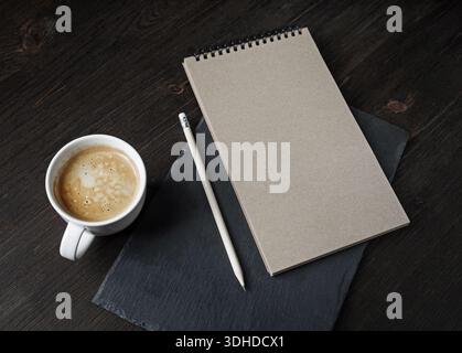 Bozzetto kraft vuoto, matita, tazza di caffè e piastra di ardesia nera su superficie in legno. Modello di branding. Foto Stock