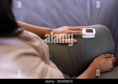Donna matura seduta sul divano che regge un dispositivo digitale bianco sulla coscia, indossa la parte superiore beige e l'anello Foto Stock