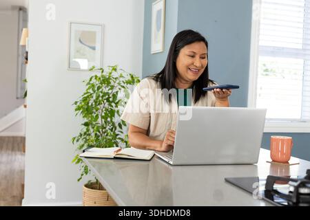 Donna asiatica matura che lavora all'isola della cucina, parla con uno smartphone mentre utilizza un computer portatile argento Foto Stock