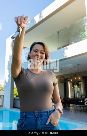 Donna afroamericana che tiene le chiavi in alto e sorridente, in piedi a bordo piscina accanto a una moderna casa a due piani Foto Stock