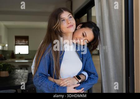 Diverse partner femminili che abbracciano e si posizionano sulla porta di casa con camicie in denim e smartwatch Foto Stock
