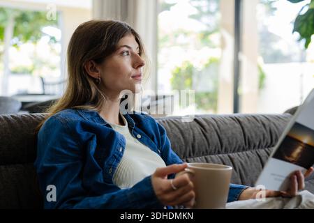 Donna seduta su un divano a casa con camicia in denim che regge tazza beige, opuscolo, sguardo all'esterno Foto Stock