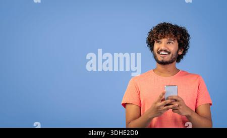 Young Man sorride mentre tiene in mano lo smartphone su sfondo blu in Studio Foto Stock
