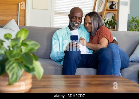 Coppie anziane diverse sedute su un divano grigio a casa, tenendo in mano lo smartphone e appoggiandosi insieme in jeans Foto Stock