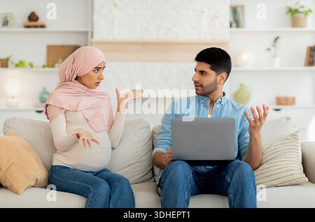 Stressata donna musulmana incinta gridando al marito mentre l'uomo usa il computer portatile, seduto insieme sul divano a casa Foto Stock