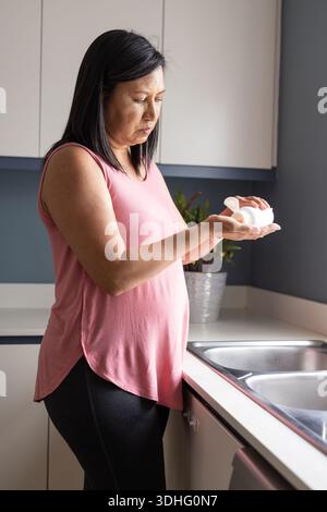Donna asiatica in cima rosa e leggings che versano pillole dalla bottiglia di pillola alla mano in cucina Foto Stock