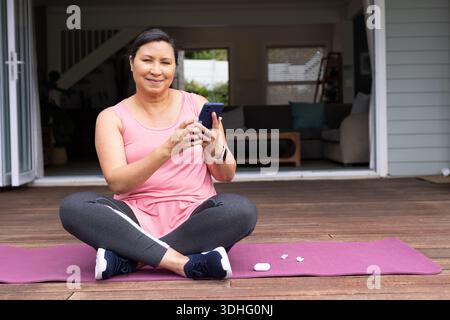 Donna asiatica matura seduta su un tappetino viola sul ponte, in mano allo smartphone, in abbigliamento sportivo con smartwatch Foto Stock