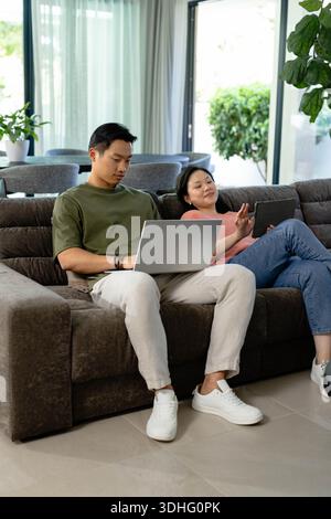 Coppia asiatica seduta sul divano marrone scuro, uomo che usa il computer portatile, donna che tiene il tablet a casa Foto Stock