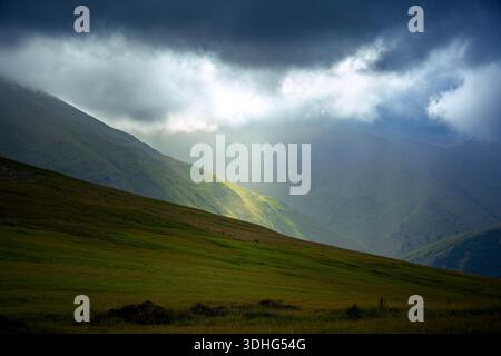 La luce del sole penetra attraverso pesanti nuvole su una valle di montagna spettacolare nel Caucaso. Atmosfera tranquilla, pendii bui e vivaci momenti salienti. 7K alto-r Foto Stock