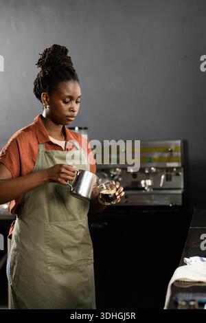 Donna africana che indossa un grembiule verde che versa latte in una tazza di vetro al bar vicino alla macchina per espresso Foto Stock