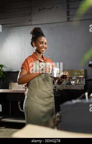 Donna adulta che sorride, prepara il caffè al bancone del bar, indossa il grembiule di oliva e tiene in mano la caraffa del latte Foto Stock