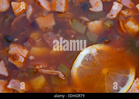 Ricca solyanka di pomodoro con fette di limone e affumicate a dadini, olive, cubetti di patate in brodo aromatico lucido, primo piano per una casa accogliente Foto Stock