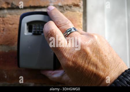 La mano di una persona che indossa un anello si vede aprire una cassetta per chiavi montata su una parete di mattoni. La cassetta portachiavi è grigia con una finitura nera e presenta una combinazione numerica Foto Stock