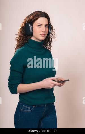Una giovane donna con capelli rossastri ricci, con cuffie e maglione verde, tiene in mano uno smartphone e ascolta la musica in uno studio. Foto Stock