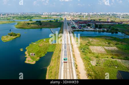 Vista aerea di un treno che attraversa un paesaggio lussureggiante, che biseca acque tranquille e conduce verso il lontano paesaggio urbano, Dacca, Dacca Divisione, Bangla Foto Stock