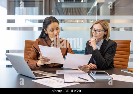 Diverse donne d'affari stanno collaborando alla creazione di documenti aziendali durante un'importante riunione aziendale in un ufficio moderno, esaminando insieme i dati finanziari e la strategia Foto Stock